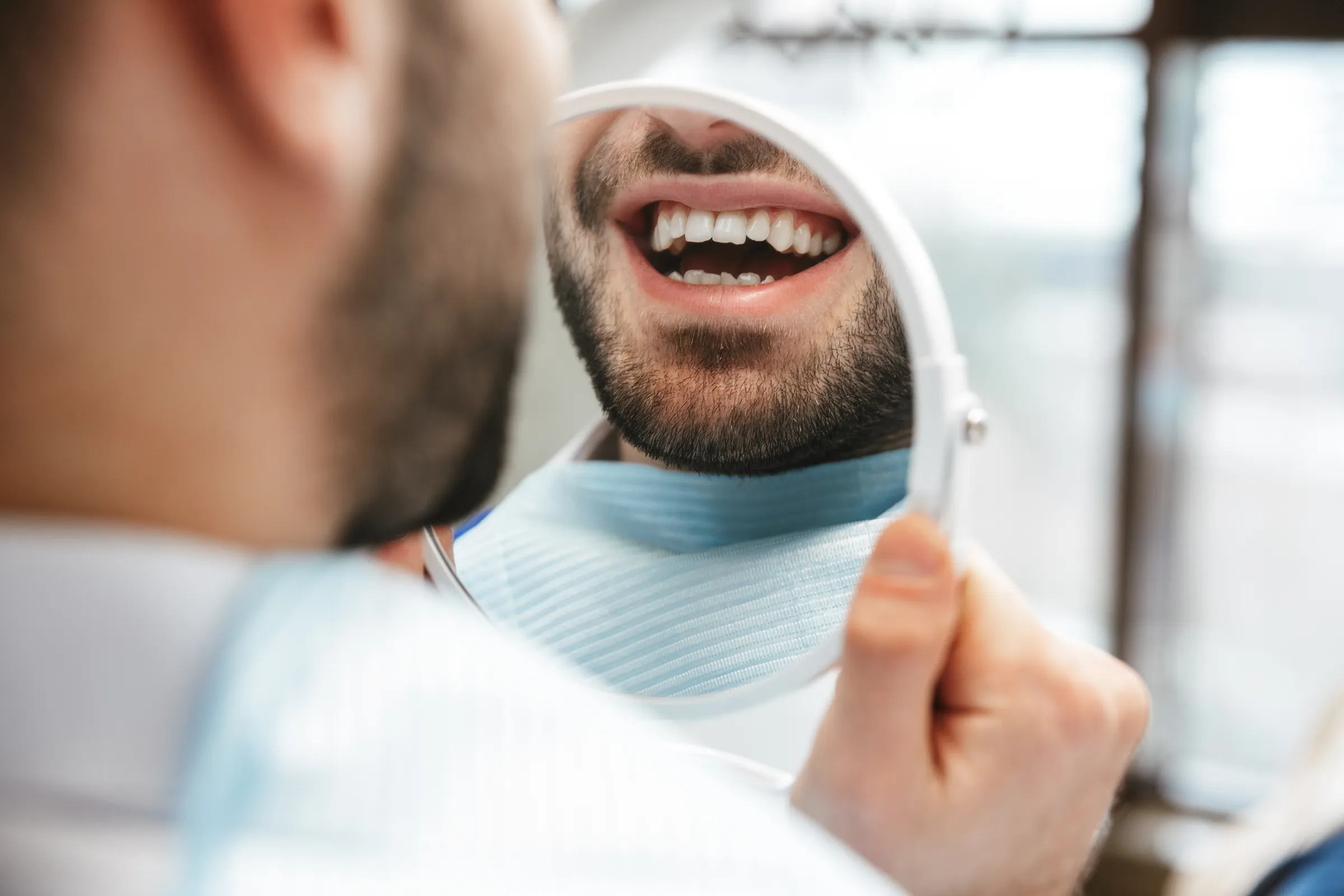Smiling man checking his teeth in a mirror held by the dentist