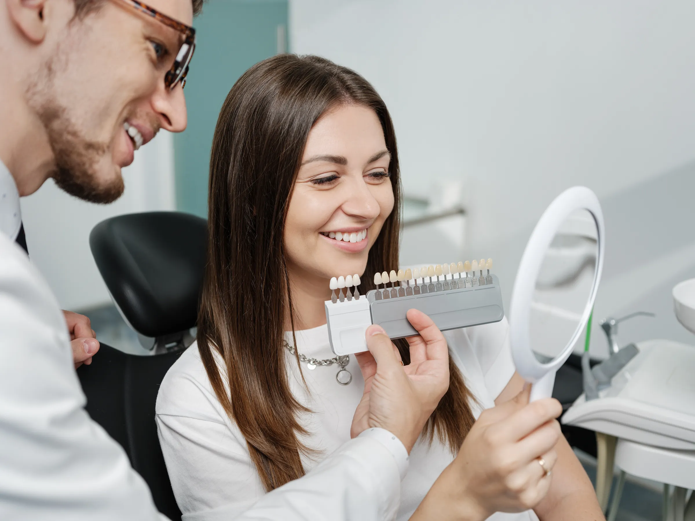 Patient comparing a shade guide to her smile in a mirror with her dentist